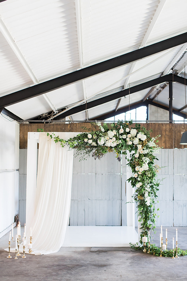 White draping with greeneries and white roses ceremony backdrop combined with gold candle holders makes a beautiful rustic-chic decor for an indoor wedding ceremony. // ❤ Check Out These Gorgeous 20 Indoor Wedding Ceremony Ideas. // http://mysweetengagement.com/indoor-wedding-ceremony-ideas