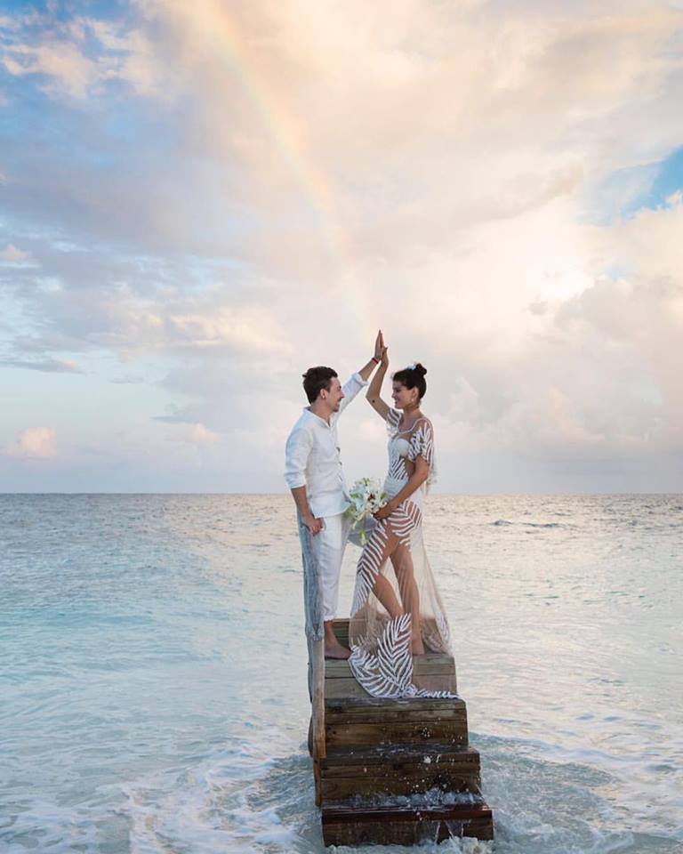 Isabeli Fontana and Di Ferrero pose holding hands high in front of the ocean with a rainbow on the background