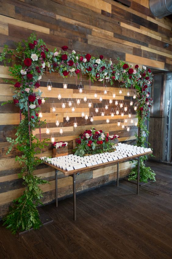Stunning escort card table setup with lots of greenery, white and deep red flowers, and magical hanging candle lights forming a beautiful backdrop. // Check out 50+ original wedding decoration ideas. // mysweetengagement.com
