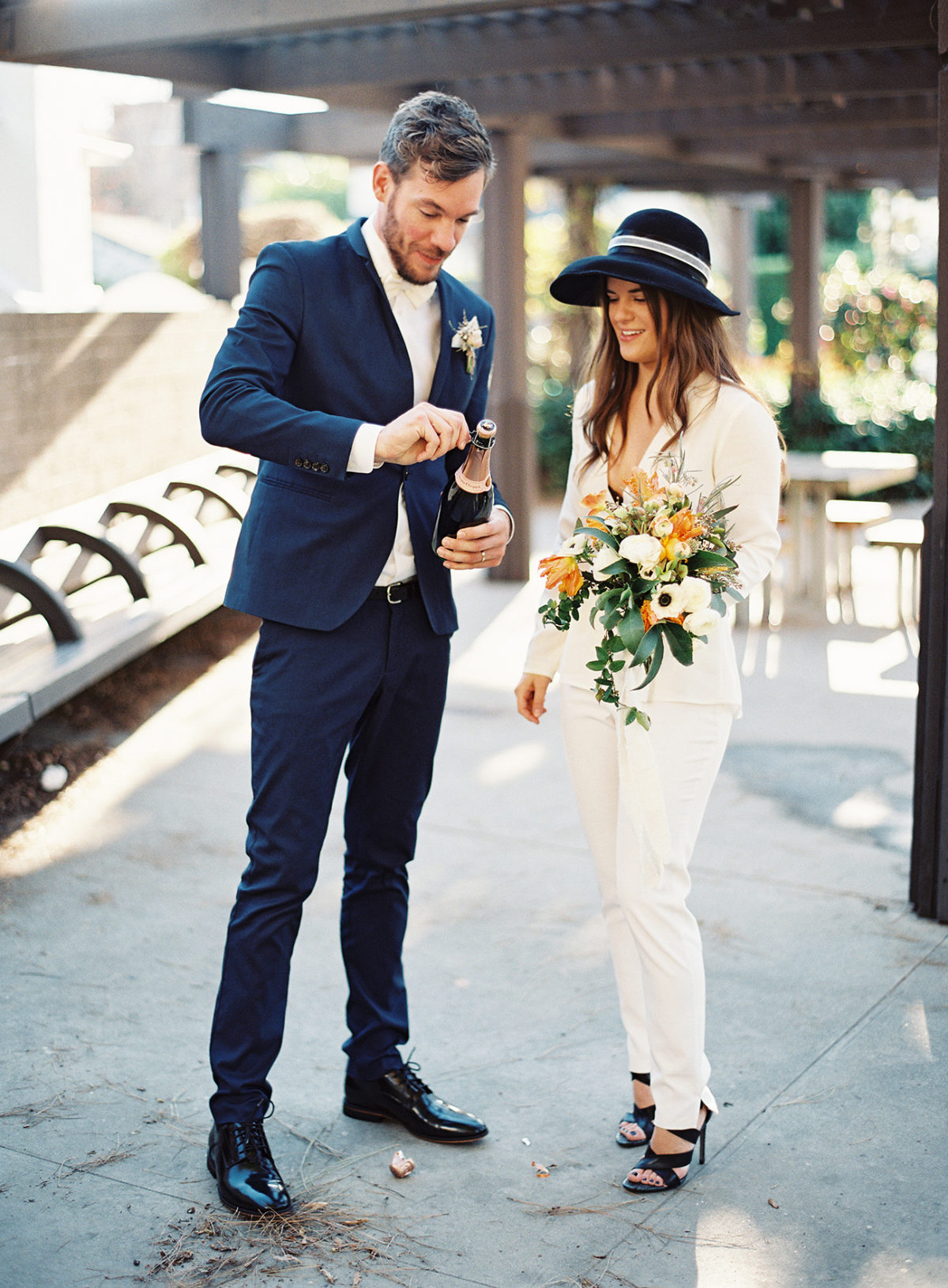 Gorgeous non-traditional modern bride and groom outfit for a courthouse celebration. Bride is wearing white blazer and pants, complimented with black heels and a statement hat. // See more: 20 Stunning Civil Wedding Outfit Ideas to Make it Official In Style. // mysweetengagement.com