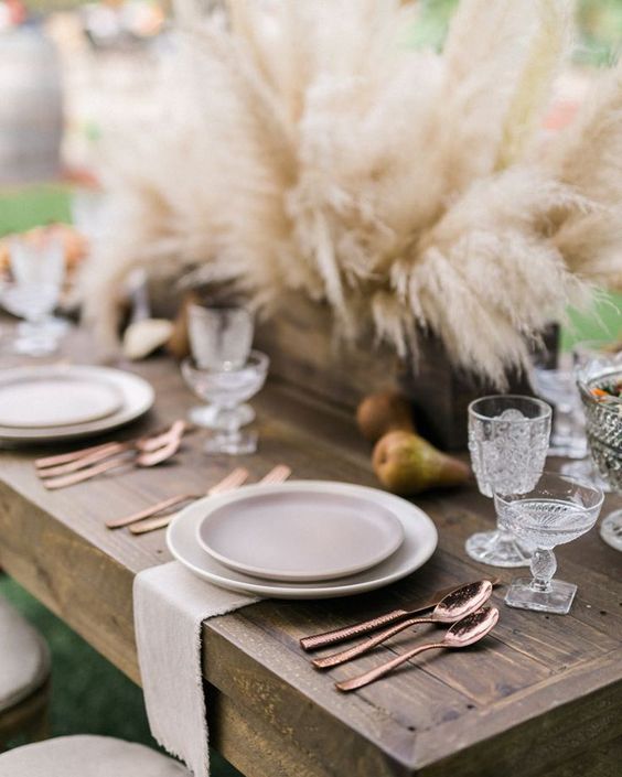 Wood table decorated with earth tone dishes, copper silverware and rustic pampas grass centerpiece.