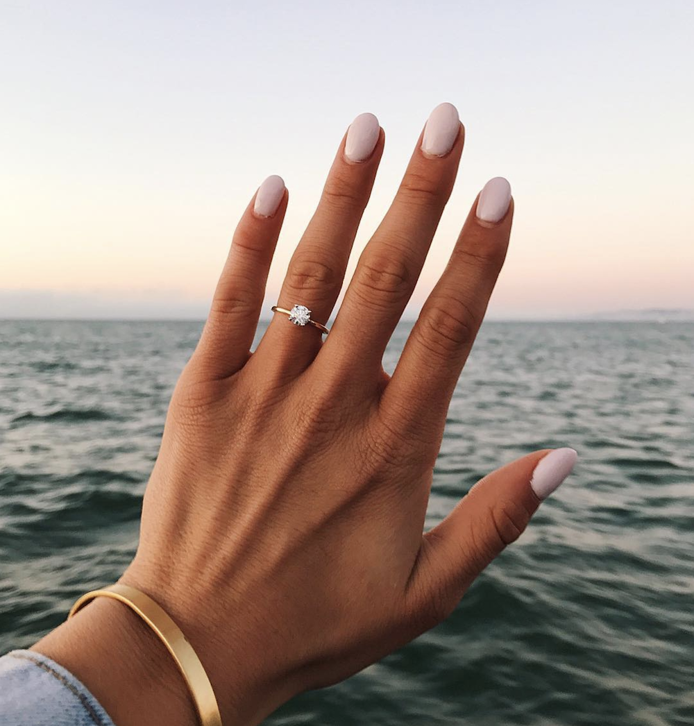 hand selfie with a solitaire gold round engagement ring with the ocean as backgroung