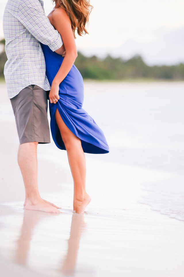 Couple hugging on a beach engagement photoshoot. Girl wears blue mid dress and guy patterned shirt with gray shorts.