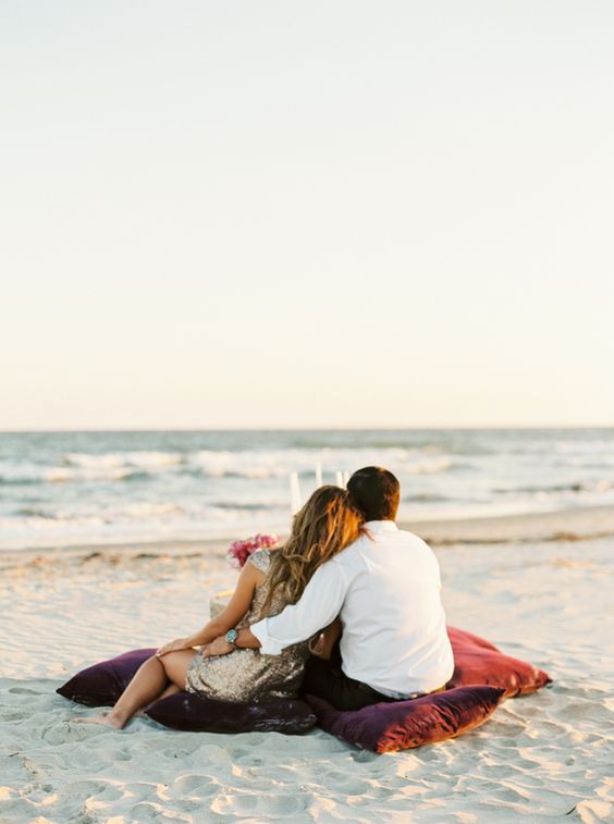 couple seating on pillows on the sand facing the ocean on a beach engagement photoshoot