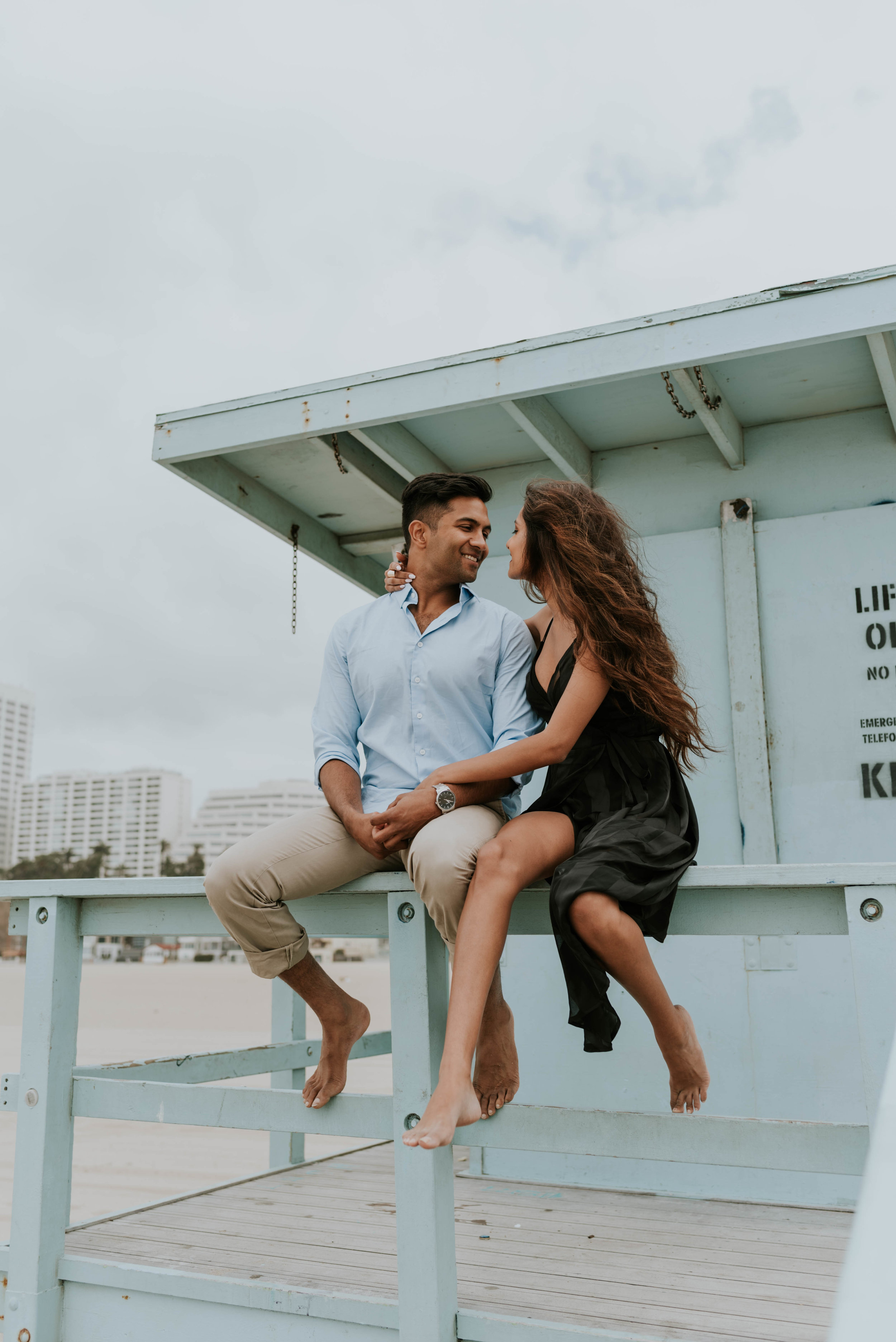 Couple on a beach engagement photoshoot. Girl wears black midi dress and guy wears blue shirt with tan pants.