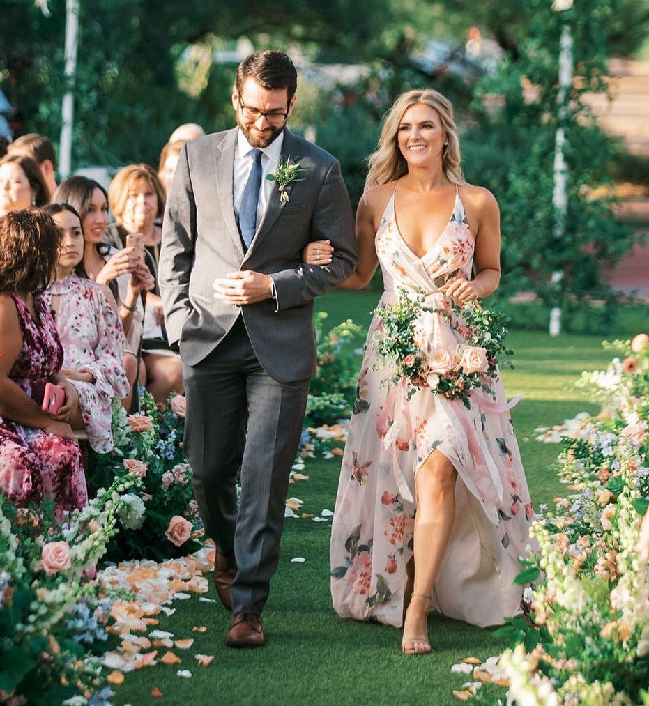 Groomsman and bridesmaid walking down the aisle. Groomsman wears gray suit and blue tie, bridesmaid wears a long blush floral dress with slit and holds a floral hoop.
