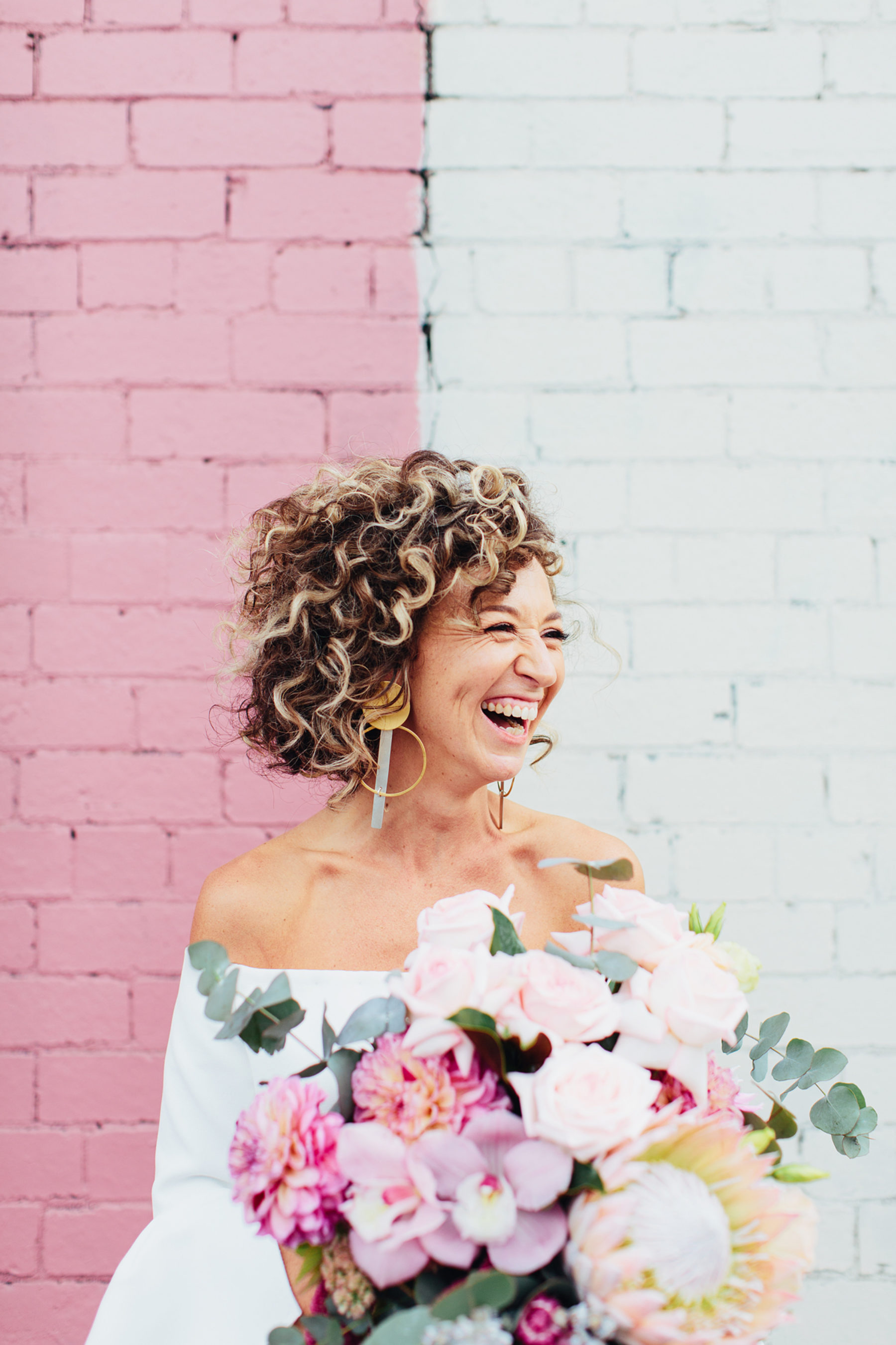 bride with blonde curly hair down and off the shoulder wedding dress and pink flowers bouquet