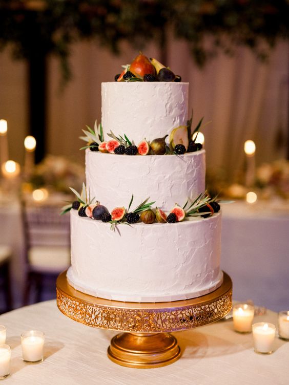 3 tiered white wedding cake design embellished with dark fruits on a gold cake stand