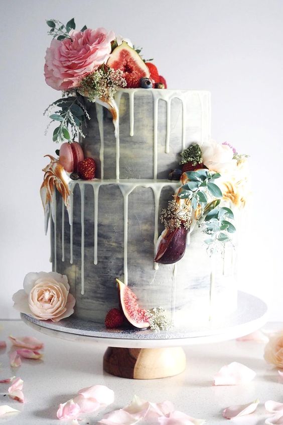 2 tiered concrete wedding cake with dripping white frosting, embellished with macaroons, pink peonies, greeneries and fruits