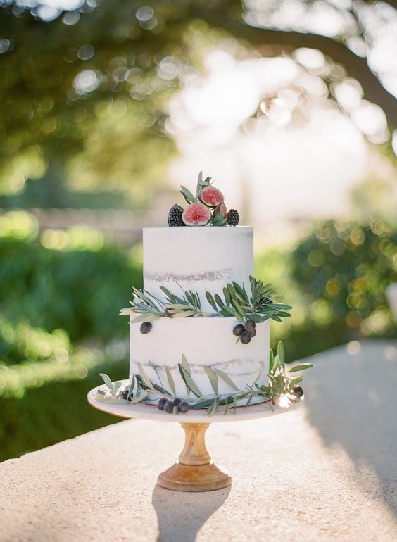 two tiered semi naked wedding cake with white frosting, decorated with greeneries, fruits and figs