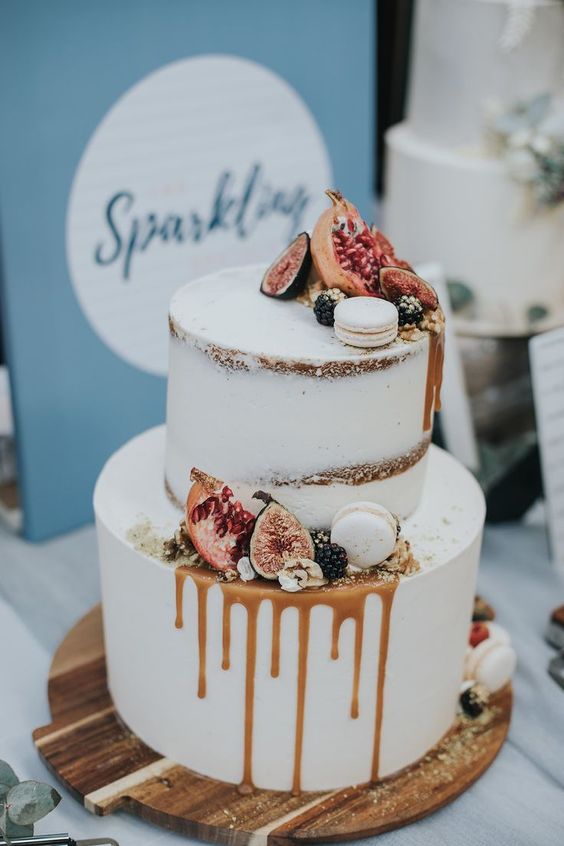 two tiered semi naked wedding cake with white frosting and caramel dripping, topped with white macaroons and autumn color fruits on a wood cake stand