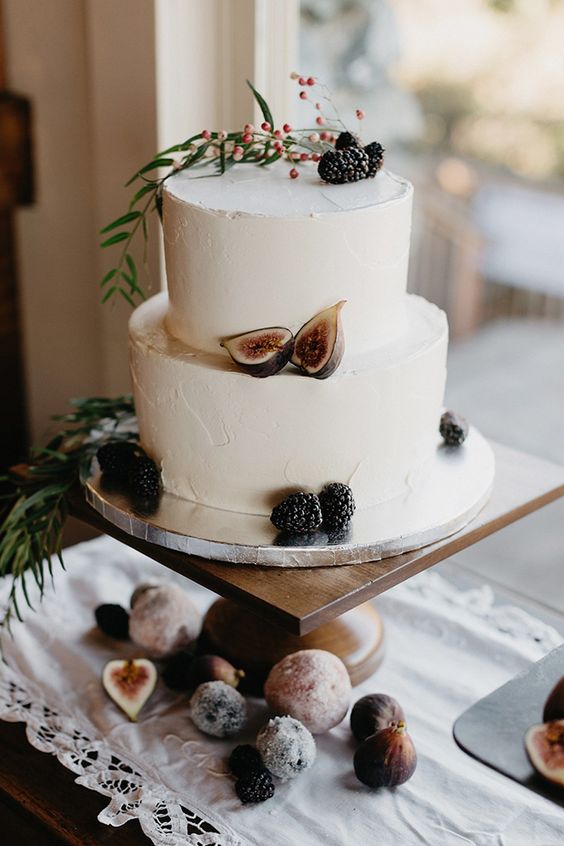 2 tier white wedding cake decorated with blackberries, figs and greeneries on a rustic wood stand