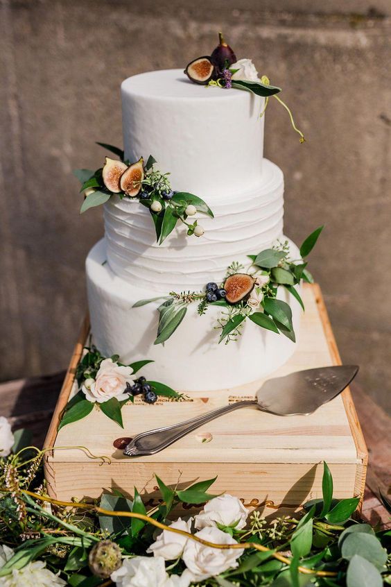 three tier white frosting wedding cake, topped with figs, white roses and greeneries on a wood stand