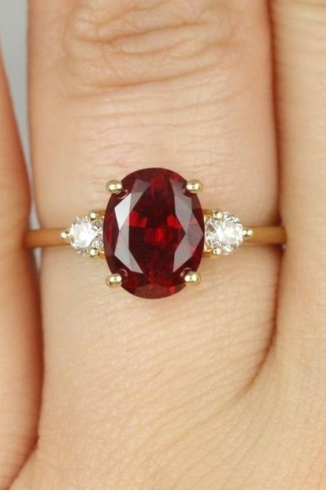 3 stone engagement ring with two round side diamonds and a center ruby oval stone on a gold band.