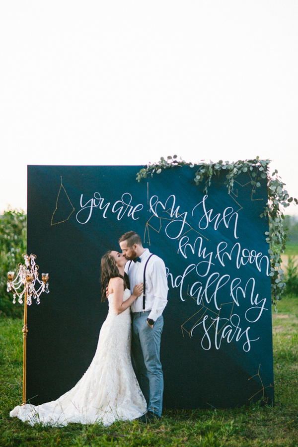 bride and groom kissing in front of a wedding ceremony backdrop with white handwritten "you are my sun, my moon and all my stars" on blue background