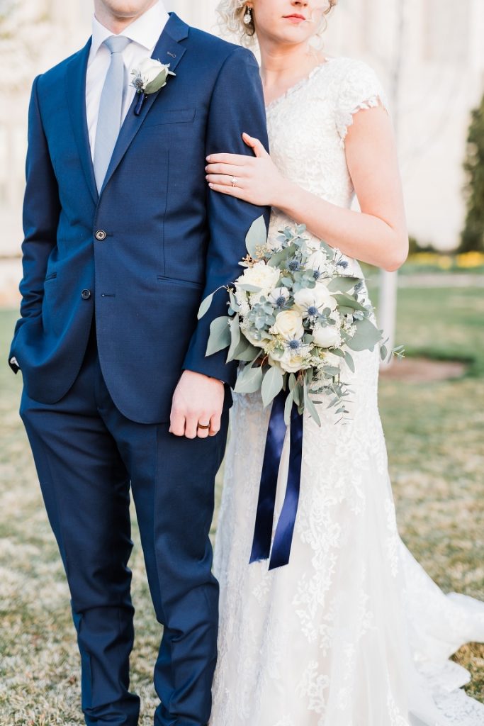groom wearing Pantone classic blue suit, dusty blue tie, white rose boutonnière and bride wearing long lace wedding dress, holding white a bouquet with white flowers, greeneries and navy blue ribbon