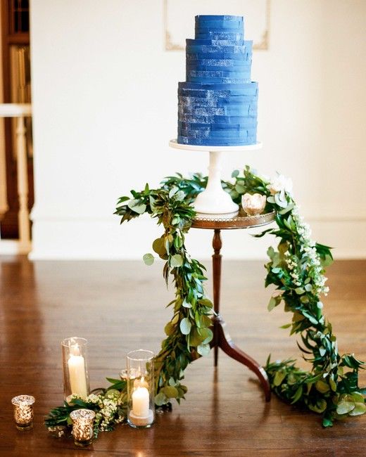 3 tiered wedding cake with layered blue frosting on a small table, with greeneries and candles decorations