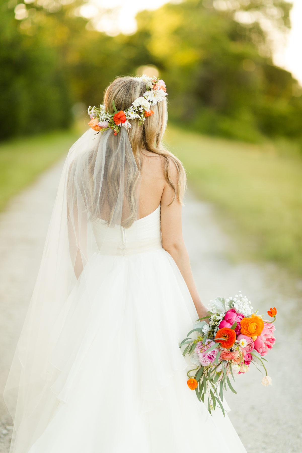 bride wearing strapless wedding dress, holding bridal pink and orange colored floral bouquet, wearing her blonde hair down, adorned with a floral crown and a long veil