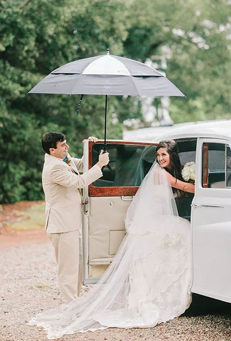 portrait of a groom holding the wedding getaway car door and an umbrella while the bride goes inside the car