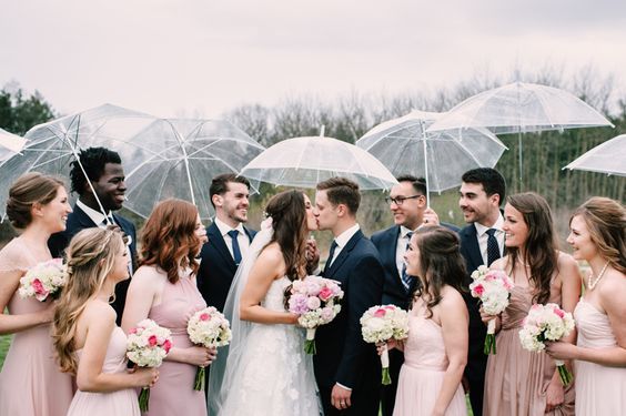 bride and groom kissing portrait holding matching clear umbrellas with their wedding party on a rainy day wedding