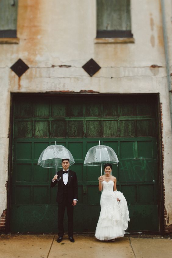 bride and groom portrait on a rainy day wedding holding clear umbrellas in front of a dark green garage door as a background
