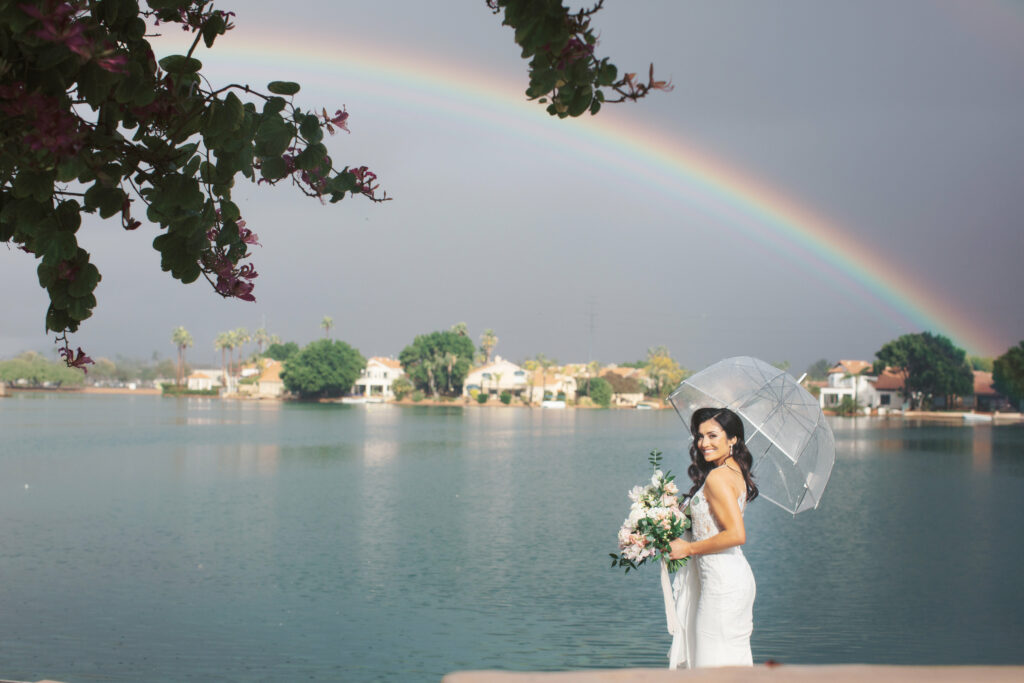 bride holding an umbrella and floral bouquet in front of a lake with gray skies and a rainbow