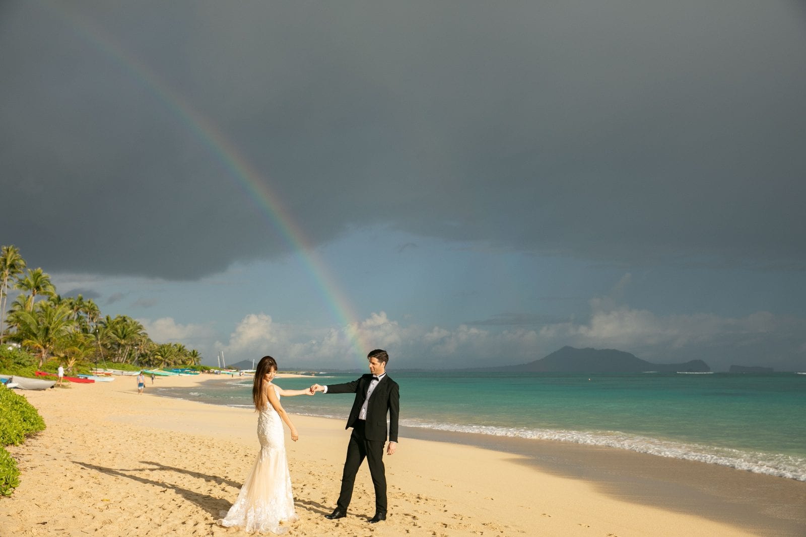 bride and groom holding hands on the beach with cloudy skies and rainbow