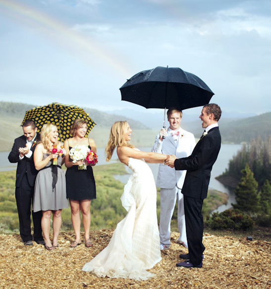 bride and groom holding hands during wedding ceremony with bridesmaids and wedding officiant holding umbrellas and a rainbow on the sky as backgrop