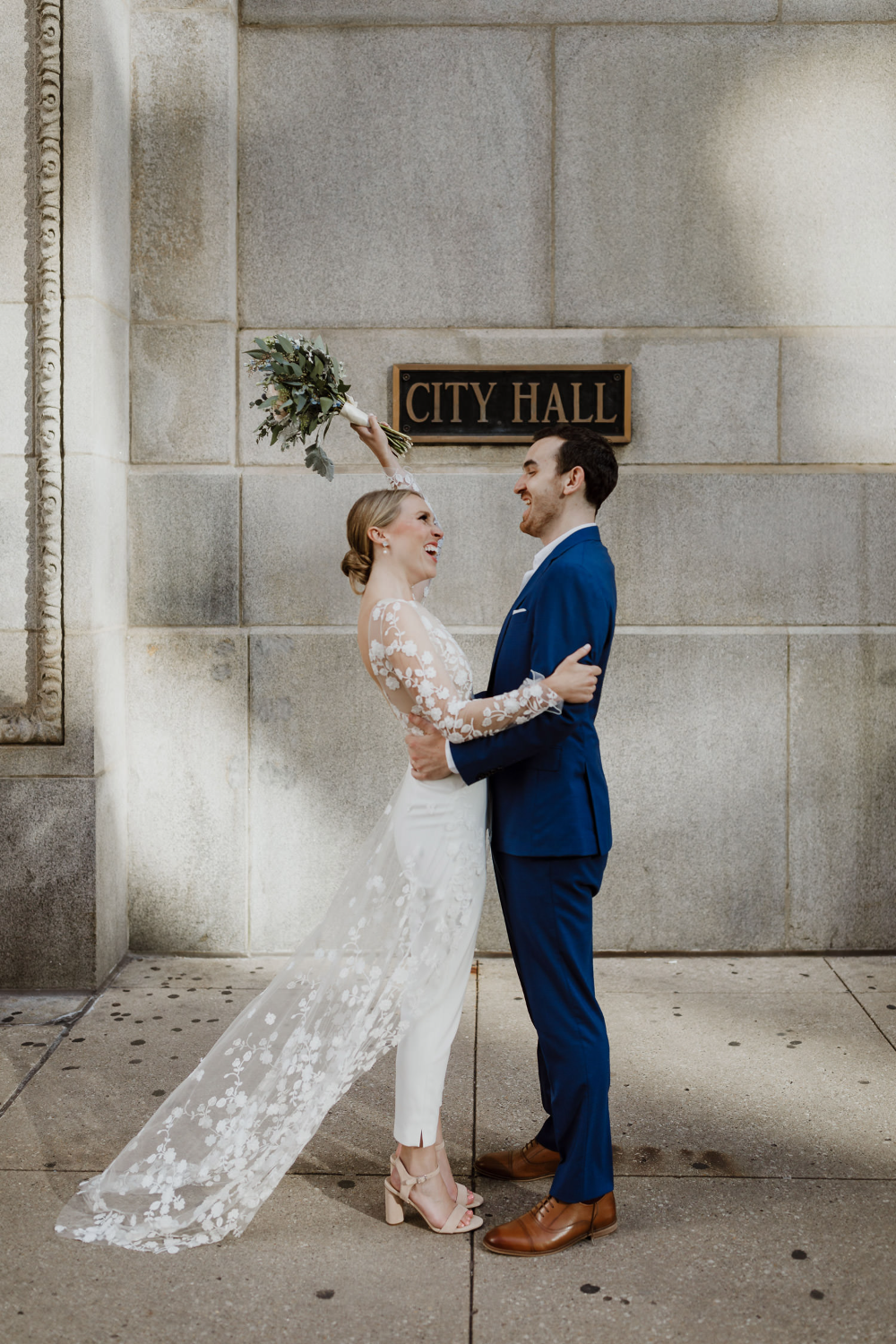 bride and groom in front of the city hall building hugging. bride holds her greenery bouquet up high