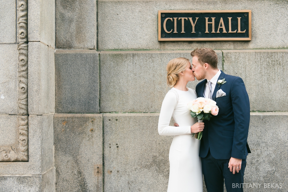 bride and groom kissing in front front of city hall sign. She wears ling sleeve dress and is holding white peonies bouquet with a low bun