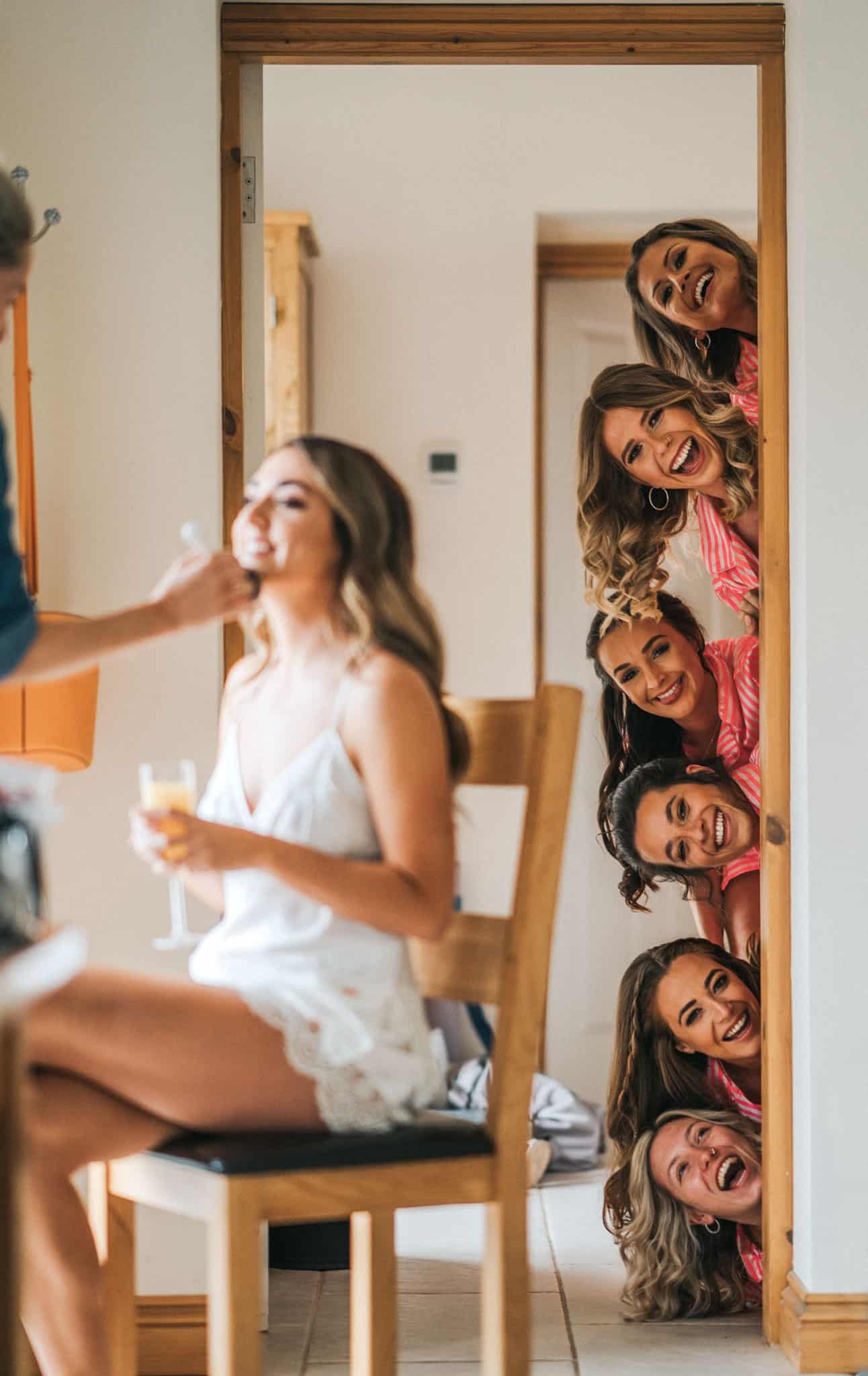 bride getting ready for her wedding day with makeup while bridesmaids take a peak at her from the door