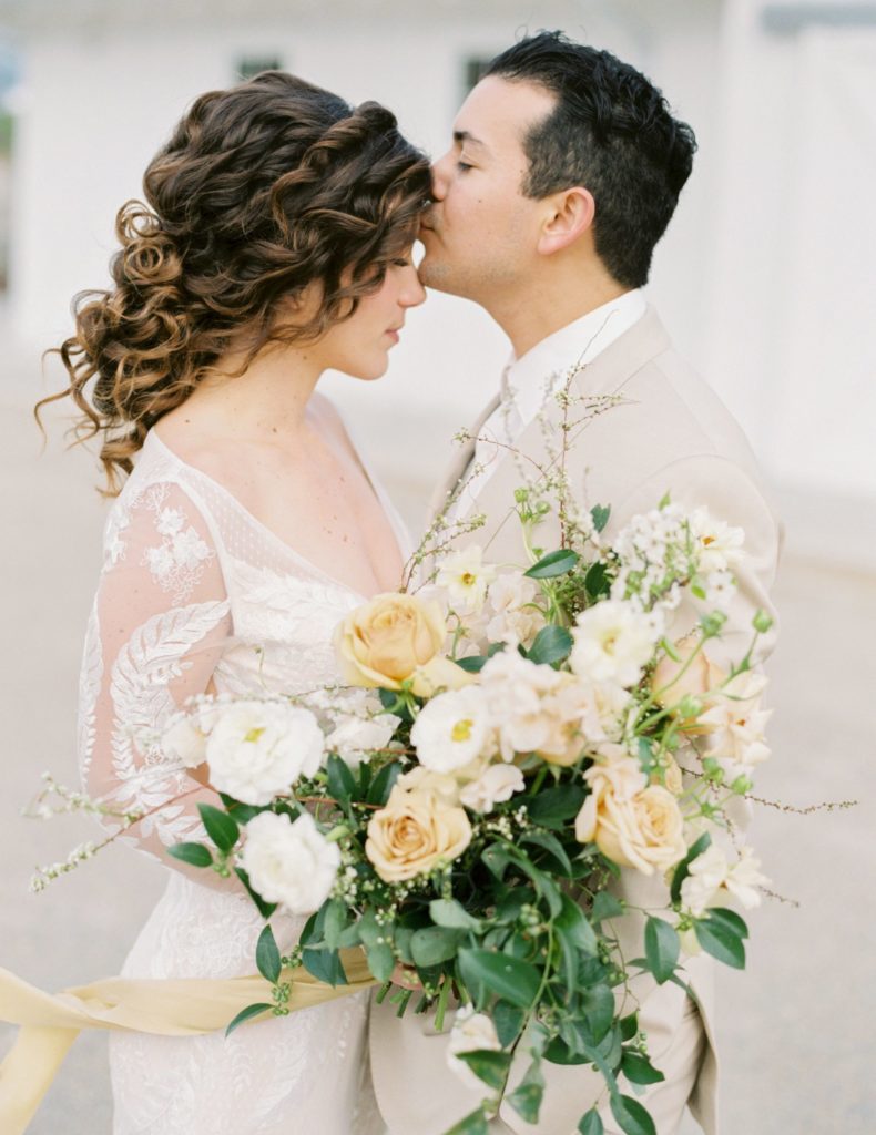 groom kissing bride's forehead. bride has curly hair and is using low messy bun and holds a big wedding bouquet with yellow and white flowers