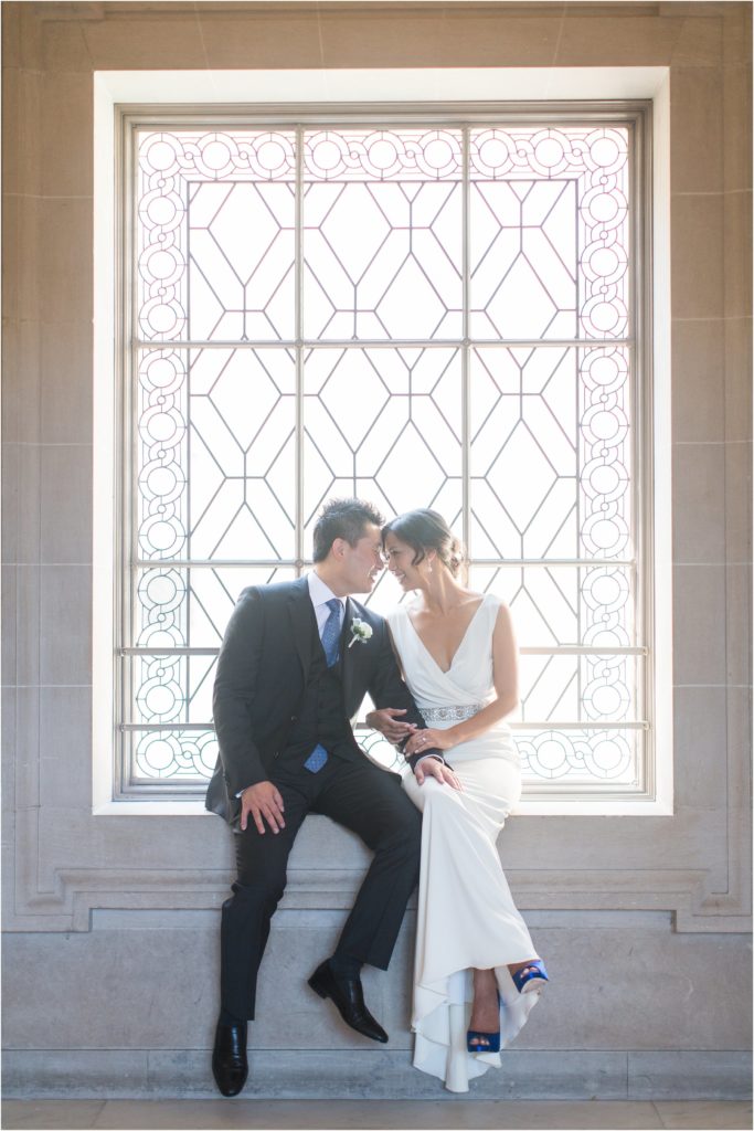 bride and groom court house wedding photo in the San Francisco city hall. The couple is sitting at the window sill head to head holding arms