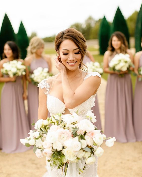 bride holding her wedding bouquet looking down with bridesmaids on the background wearing a long blush pink dress