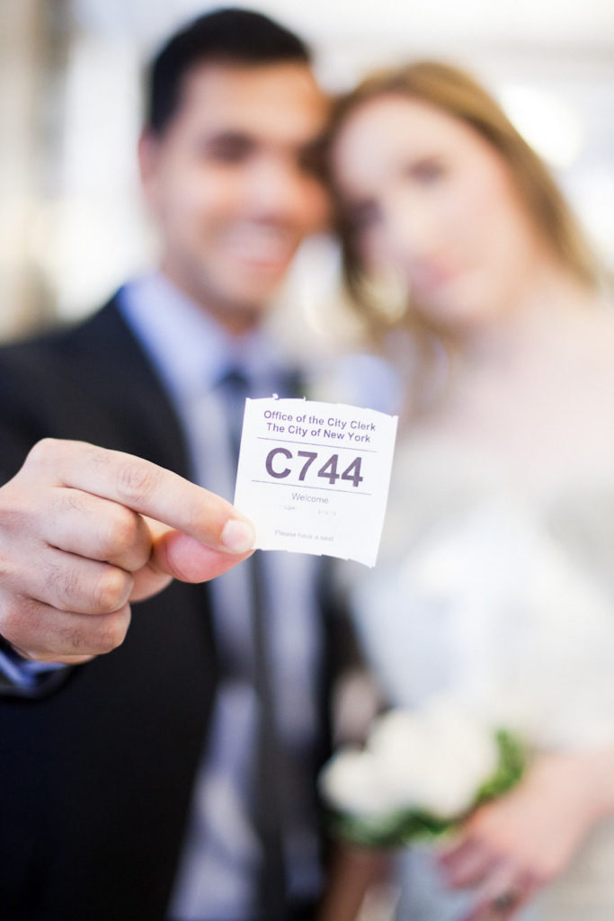 bride and groom blurred on the background holding an office of the city clerk of the city of New York ticket on the foreground with their wait number for civil marriage