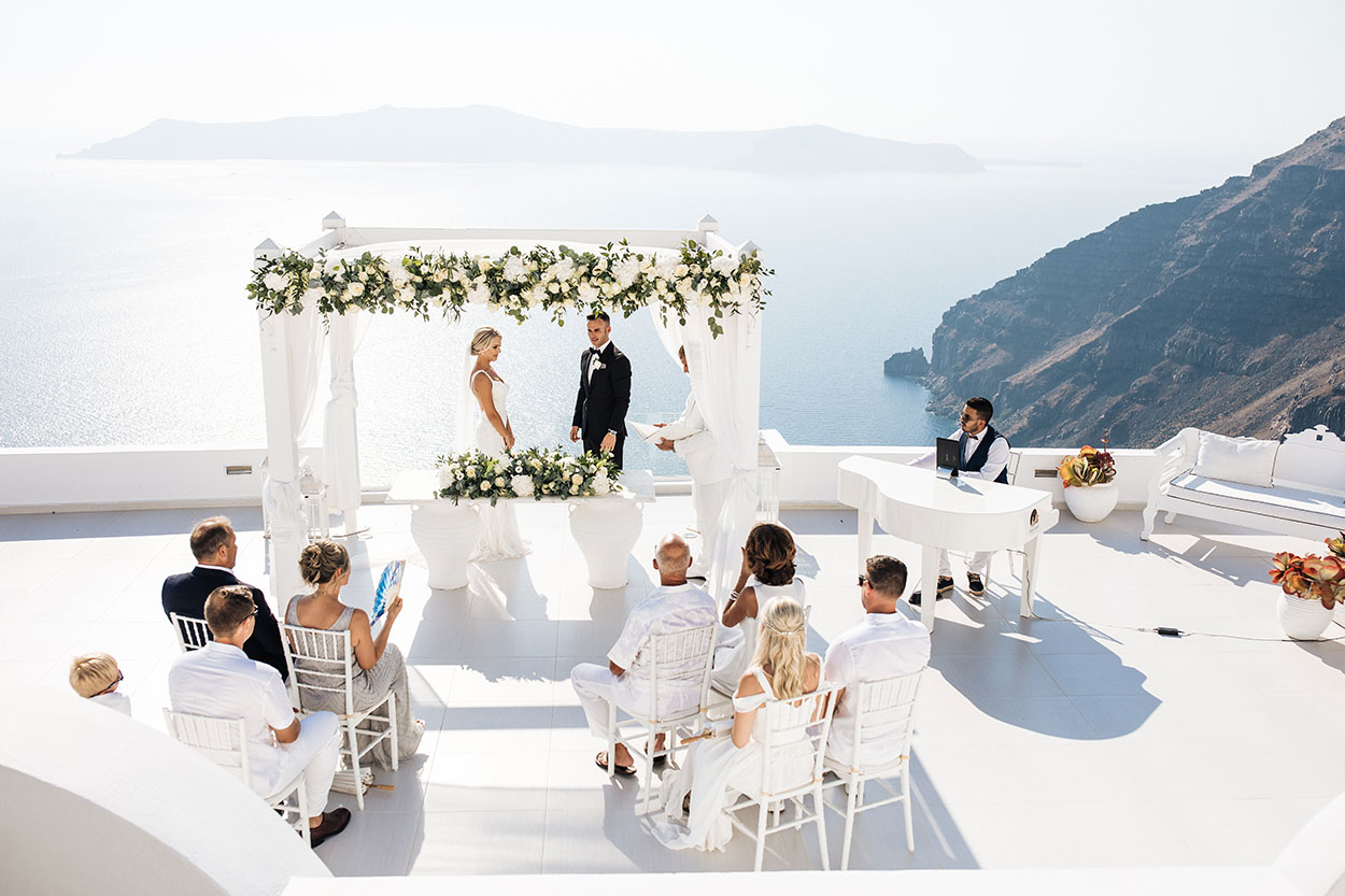 Intimate Destination in Santorini. Waterfront wedding ceremony with bride and groom under the altar decorated with white flowers and draperies. All guests wear white.